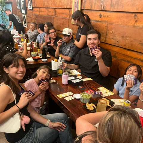 Group of people sitting around a wooden table in a casual dining setting, enjoying drinks and food.