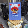 Card with cartoon character and text 'Fire Juice' on a s'mores stack with a knife, compass, and pepper grinder in the background.