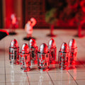 A group of metallic bullet-shaped game pieces with printed numbers, arranged on a glossy tiled surface under dramatic red lighting. A red and black textured object is partially visible on the left, with a blurred indoor background.