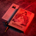 A book-style product titled "Labyrinth" for an adventure game, shown open on a textured surface under dramatic red lighting, with a printed insert card and a small die beside it.