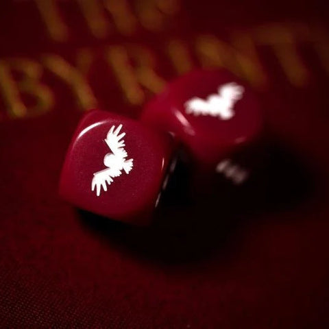 Two red polyhedral dice with white bat silhouettes, resting on a dark red textured surface with gold lettering in the background.