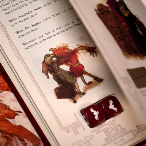 An open book or rulebook showing printed fantasy artwork and text, with two dark red dice placed on the page. The scene is photographed at an angle with shallow depth of field, focusing on the dice and nearby illustration.