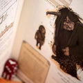 An open book or art print showing fantasy creature illustrations, with a dark horned figure and smaller character sketches on aged paper. A red die is visible in the blurred foreground.