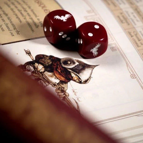 Two red polyhedral dice with white bat illustrations resting on an illustrated game manual, with a blurred brown foreground element in the lower left.