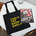 Black tote bag with 'Table Top Book Shelf' text on a white surface with a plant and book.
