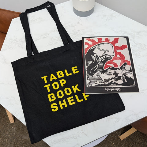 Black tote bag with 'Table Top Book Shelf' text on a white surface with a plant and book.