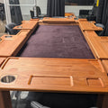 A large wooden game table with integrated side tables and cup holders, featuring a dark purple felt playing surface, surrounded by black chairs in an office setting.