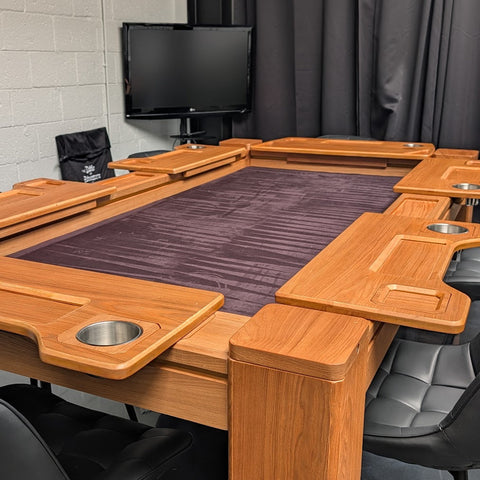 A wooden gaming table with integrated side trays and cup holders, a dark purple felt playing surface, and black chairs in a modern room setting.