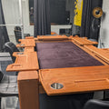 A wooden automatic slot machine table with a dark purple felt playing surface, set in a modern room with black chairs and glass windows.