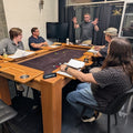 A group of people gathered around a wooden table with a dark playing surface in a modern indoor room with large windows and dark walls.