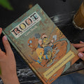 Person holding a 'Root' board game box on a table with a cup of coffee.