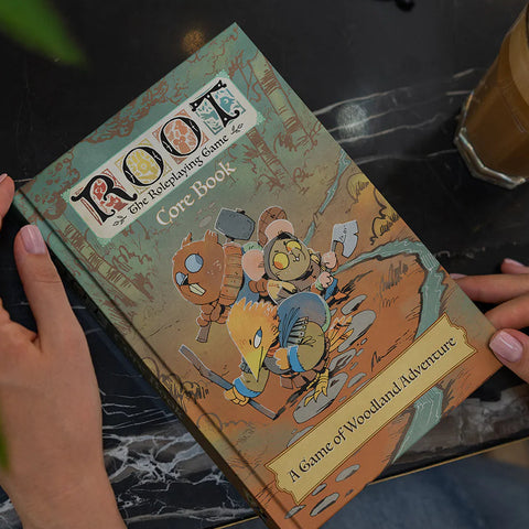 Person holding a 'Root' board game box on a table with a cup of coffee.
